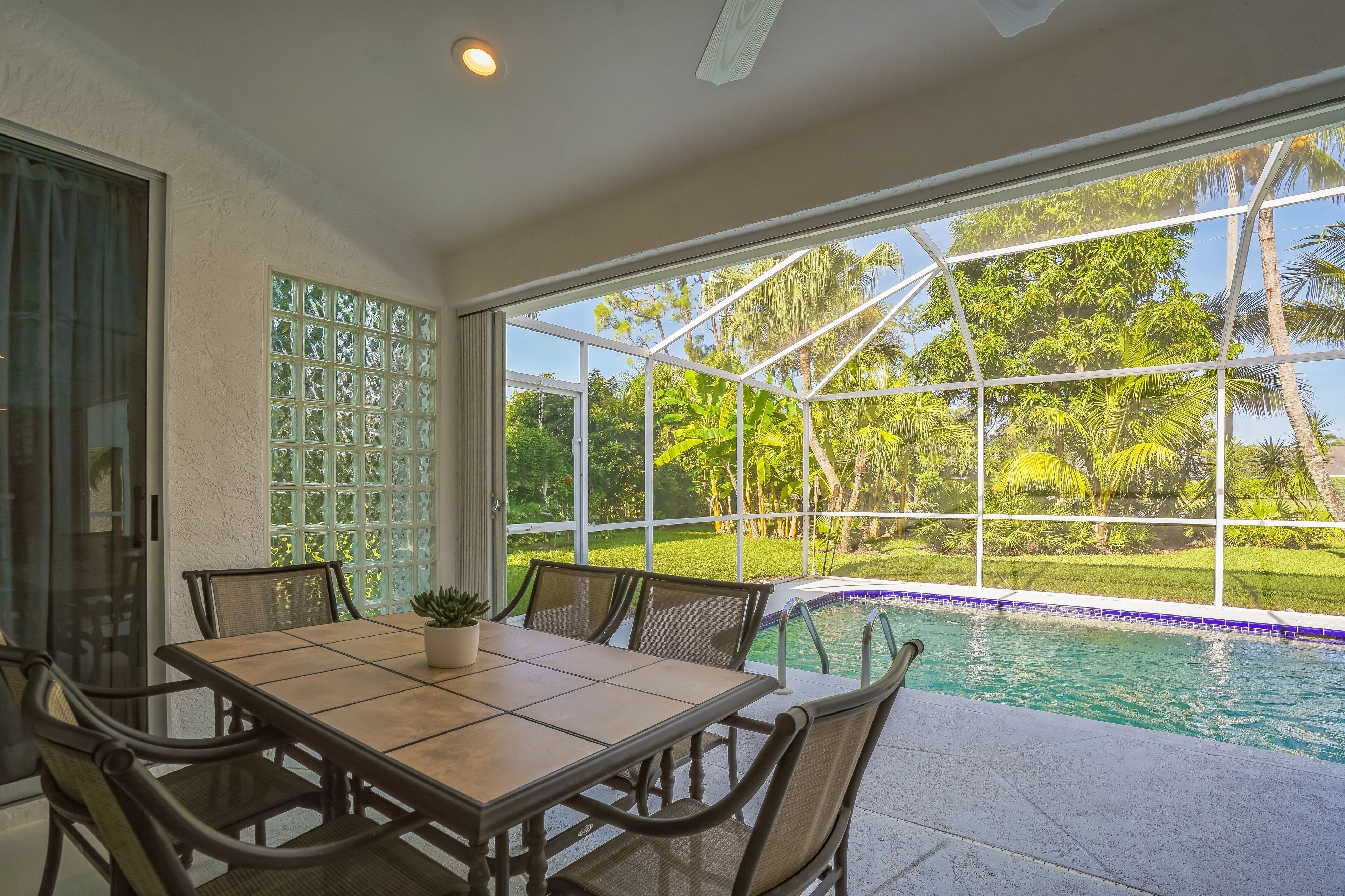 Un comedor con mesa y sillas, una planta en maceta, y vistas a una piscina y jardín tropical a través de amplios ventanales.