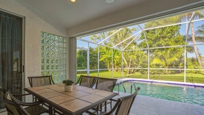Un coin repas avec une table et des chaises, une plante en pot, et une vue sur une piscine et un jardin tropical, le tout visible à travers de grandes fenêtres.