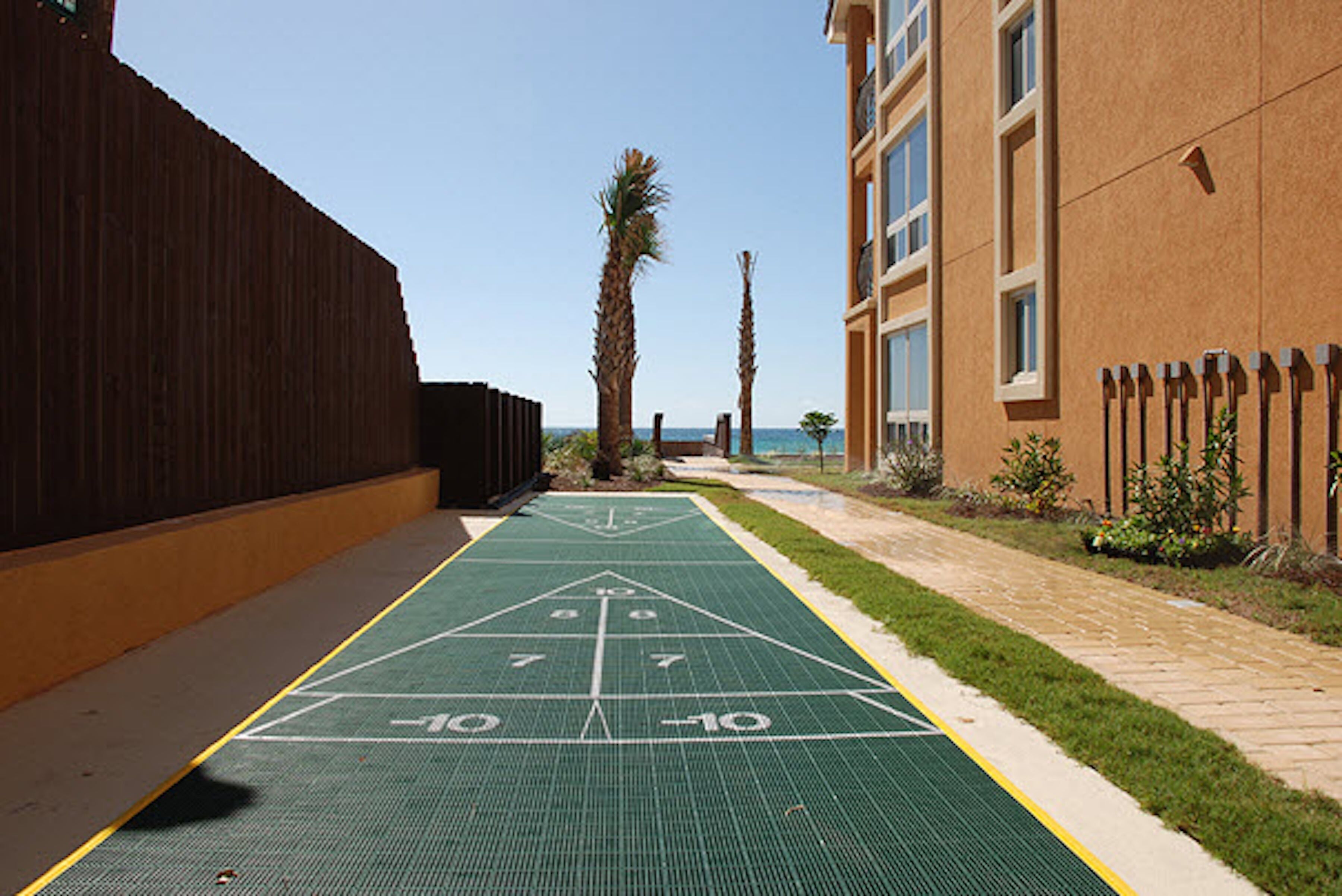 A green shuffleboard court adjacent to a building with a brick facade and a wooden fence.