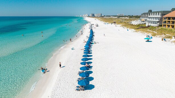 A beach with a row of blue umbrellas and beach chairs, a sandy shore, and buildings in the background. - Coral Reef Club 36 3 Bedroom Condo (Destin)