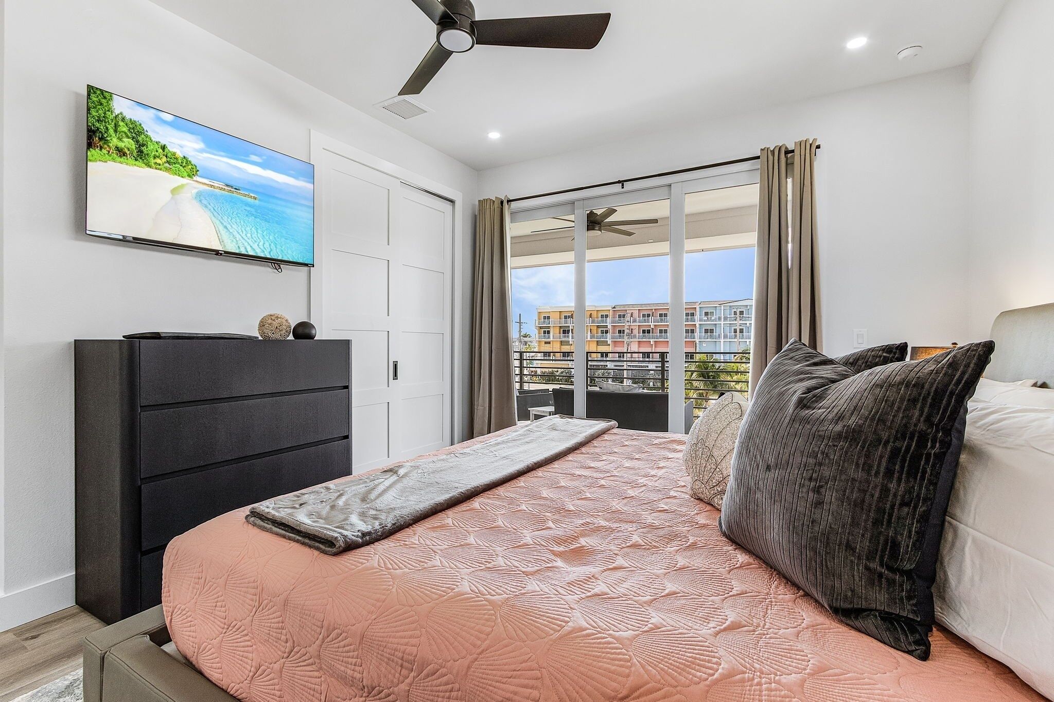 A bedroom with a bed, a TV on the wall, a ceiling fan, and a view of buildings through the window.