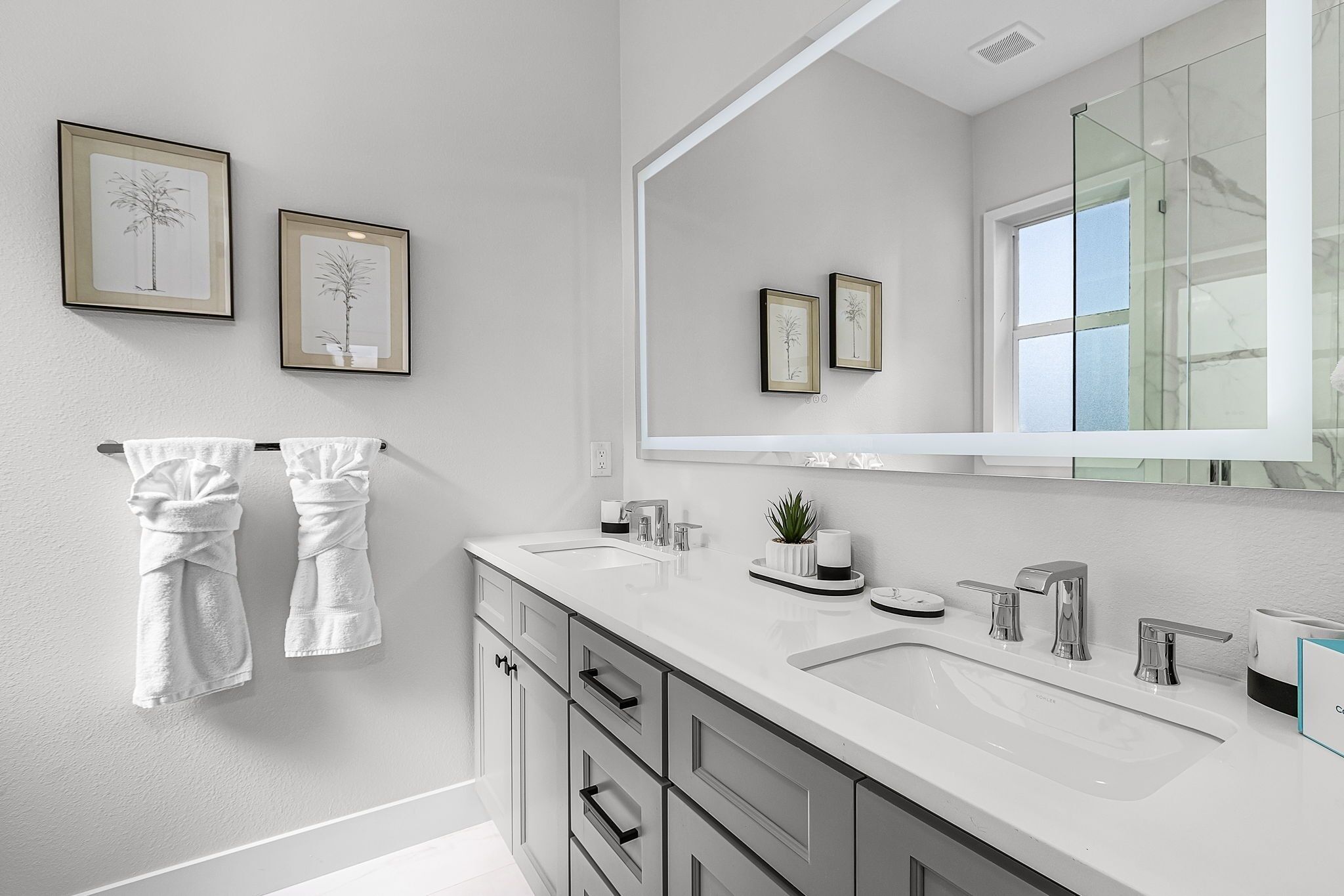 A modern bathroom with two sinks, a large mirror, and framed botanical prints on the wall.