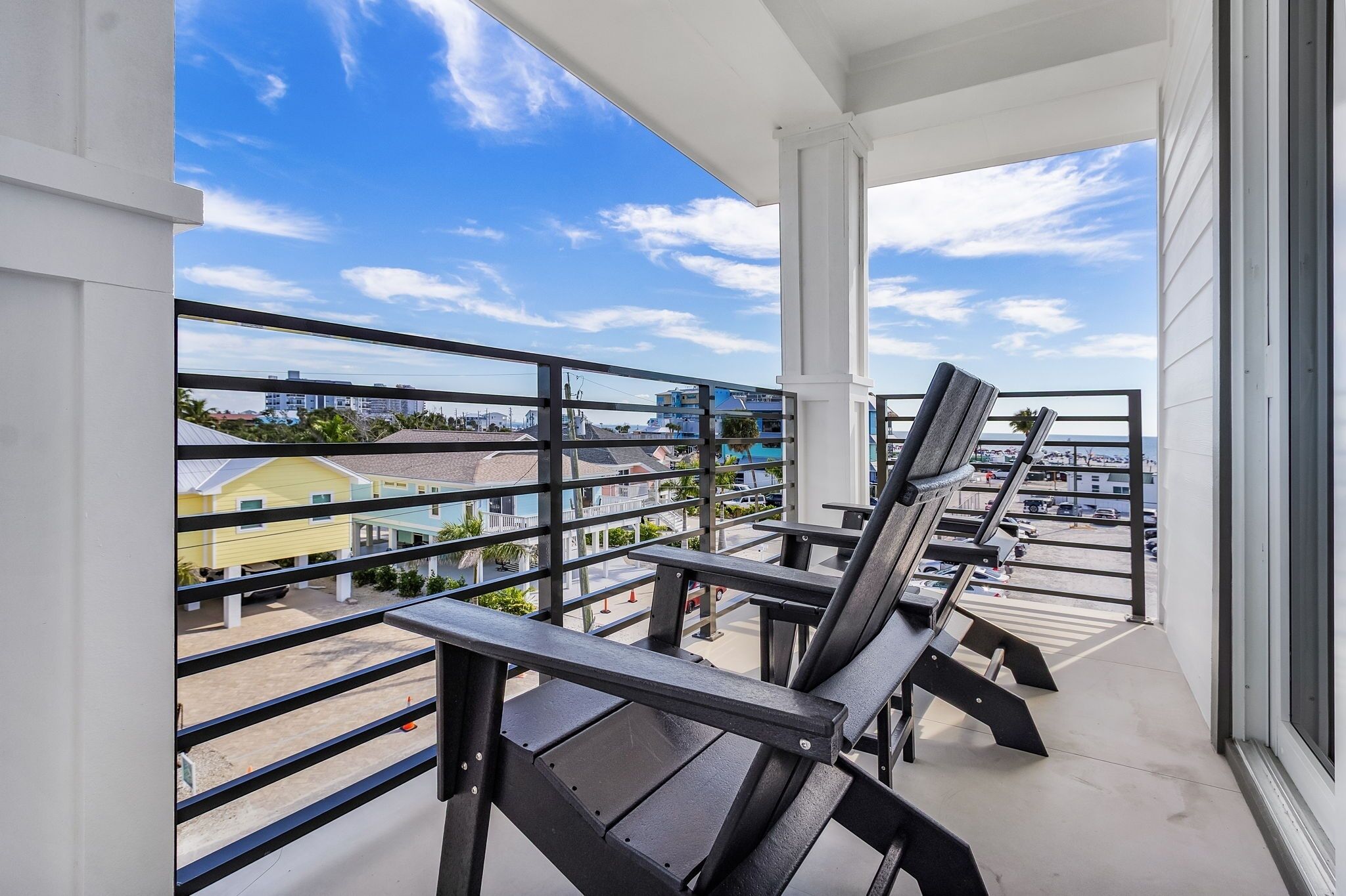 A balcony with two black chairs and a table, overlooking a parking area and buildings.