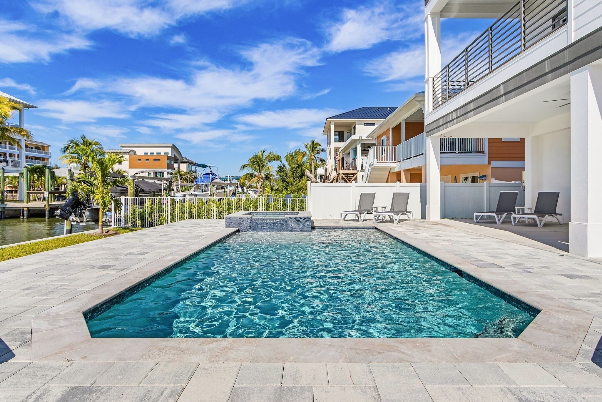 A rectangular swimming pool with a tiled deck, surrounded by buildings and palm trees.