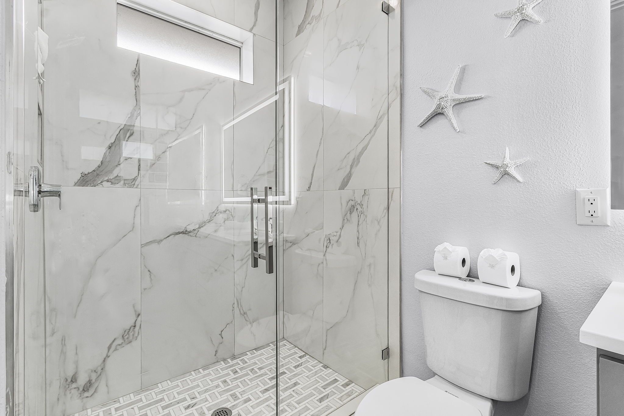 A modern bathroom with a glass-enclosed shower, marble walls, a toilet with toilet paper, and starfish wall decor.
