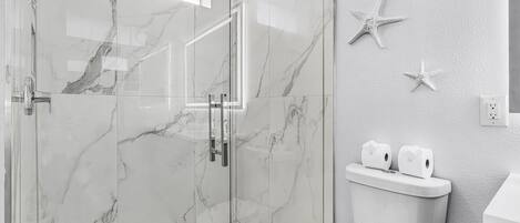 A modern bathroom with a glass-enclosed shower, marble walls, a toilet with toilet paper, and starfish wall decor.