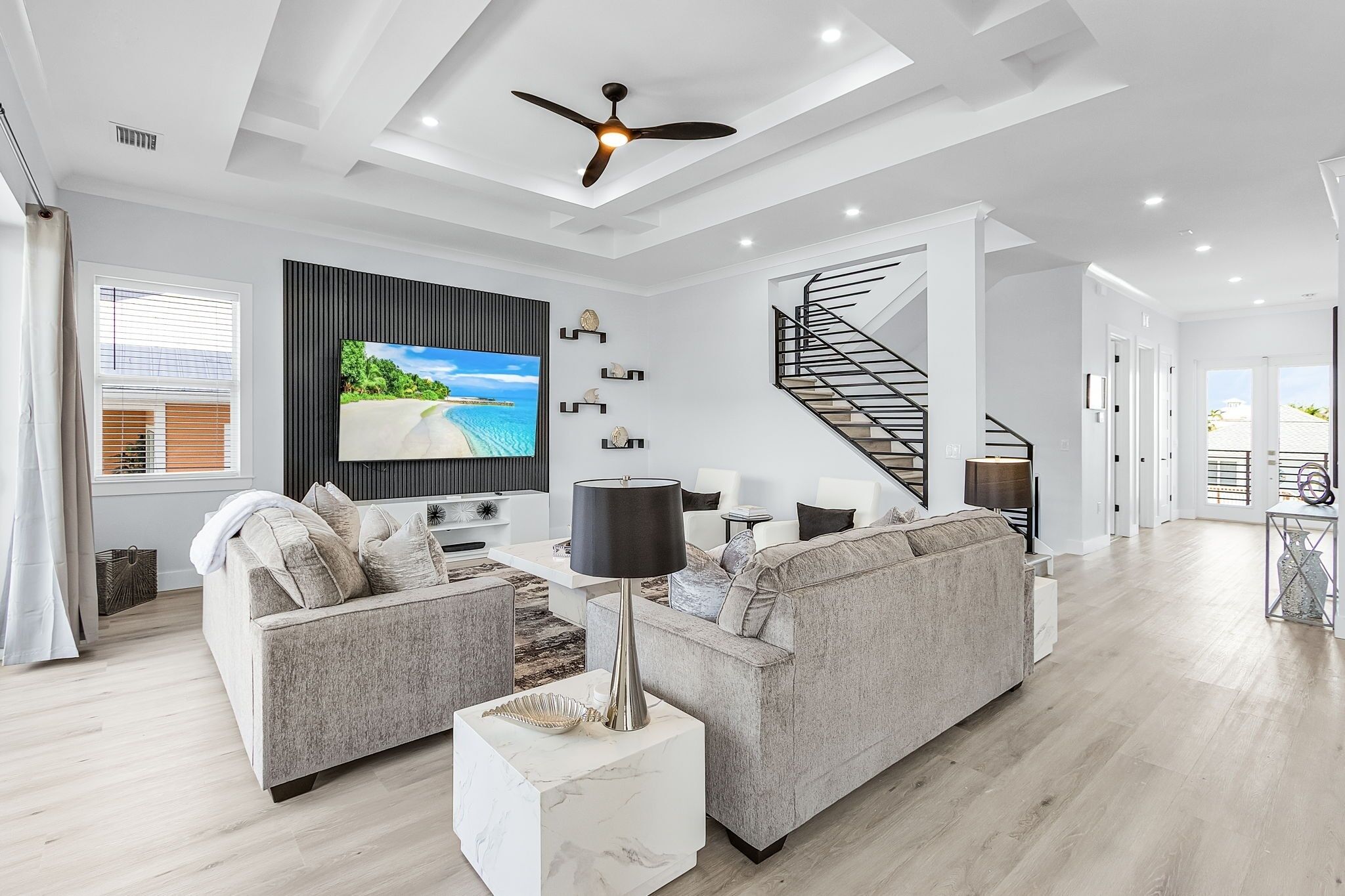 A modern living room with a grey sofa, a TV mounted on the wall, a black ceiling fan, and a spiral staircase.