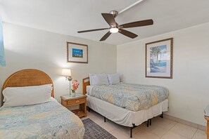 Ein Hotelzimmer mit zwei Einzelbetten, einem Deckenventilator, einem Nachttisch mit Lampe und Blumen sowie zwei gerahmten Bildern an der Wand.