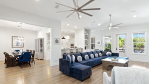 A spacious living room with a large blue sectional sofa, a dining area with a wooden table and chairs, and a kitchen in the background.