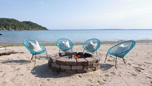 A beach with a fire pit and three blue chairs with white cushions. - Traverse City Lakefront: Beach, Dock & Hot Tub! (Traverse City)