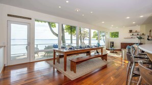 A spacious dining area with a wooden table and benches, large windows offering a view of the water, and a kitchen area with bar stools. - Traverse City Lakefront: Beach, Dock & Hot Tub! (Traverse City)