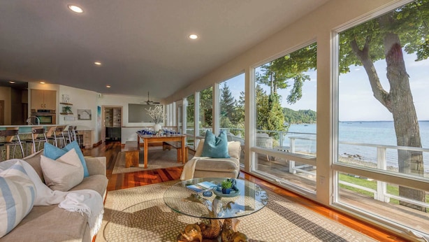 A spacious living room with a large glass coffee table, a sofa with cushions, and a view of the water through large windows. - Traverse City Lakefront: Beach, Dock & Hot Tub! (Traverse City)