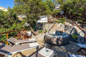 Een terras van natuursteen met lounge fauteuils, een jacuzzi en een trap, omgeven door bomen.