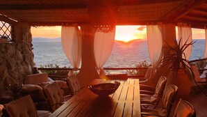 A wooden table with chairs and a bowl, set against a backdrop of a sunset over the sea, with a stone wall and a plant on the right.