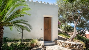 A white building with a wooden door, surrounded by greenery and a stone wall.
