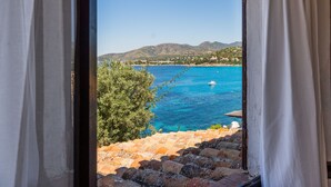 A coastal view through a window with curtains, featuring a clear blue sea, a boat, and distant hills.