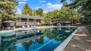 Un área de piscina moderna con sillones de descanso y sombrillas, rodeada de árboles y edificios.