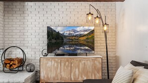 Una sala de estar moderna con un televisor de pantalla plana montado en una pared de ladrillo blanco, un mueble de madera debajo y un rincón de estar acogedor con un sofá y almohadas.