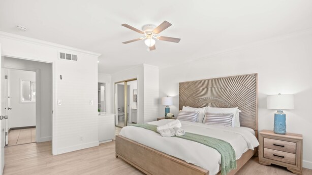Un dormitorio con cama de madera, paredes blancas, ventilador de techo y mesita de noche con lámpara.