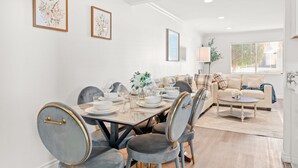 Un comedor con una mesa dispuesta para cuatro, rodeada de sillas grises tapizadas con detalles dorados. Se ve un sofá y una mesa de centro al fondo, y las paredes son blancas con una ventana que tiene persianas.