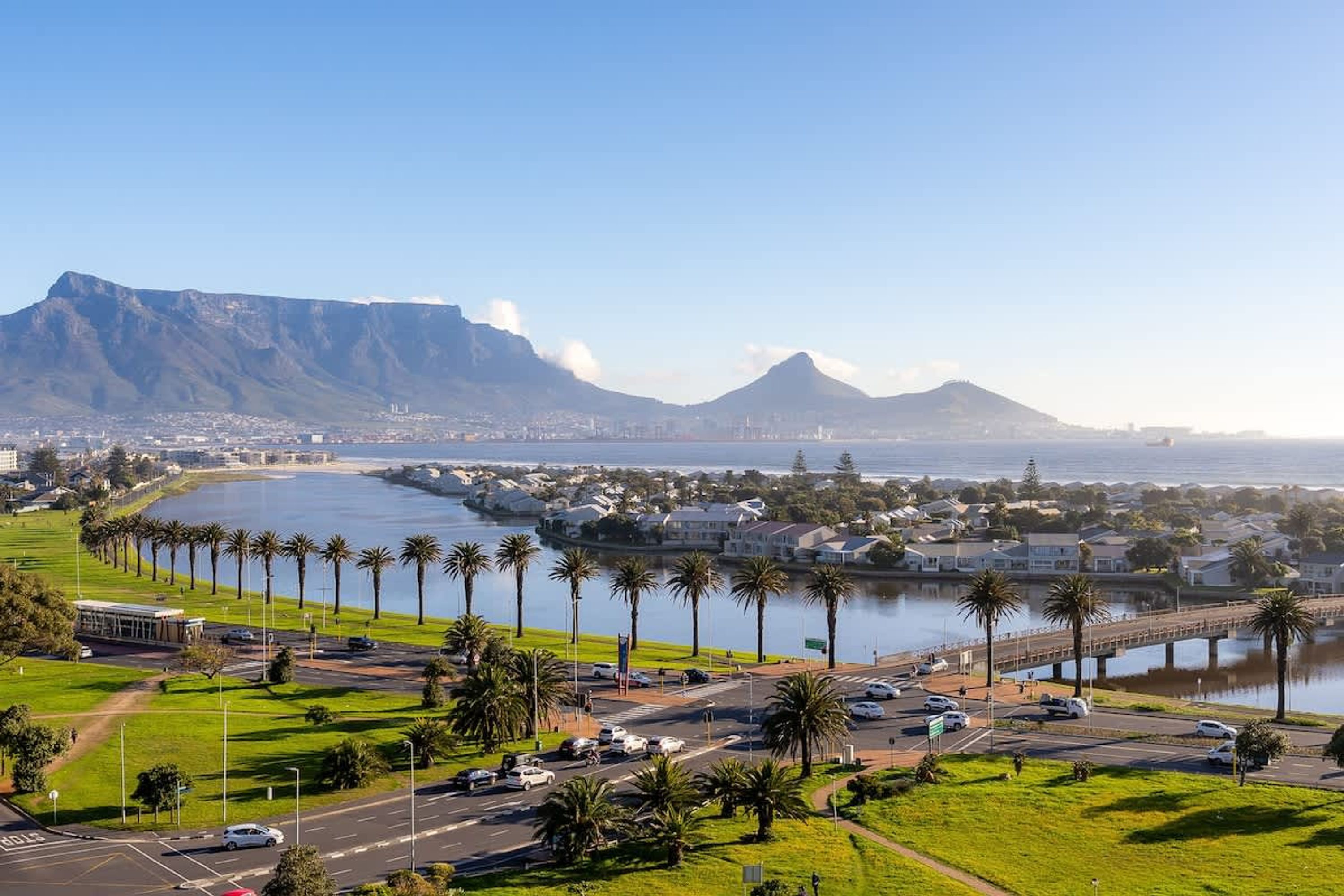A coastal cityscape with a marina, palm-lined promenade, and mountains in the background.