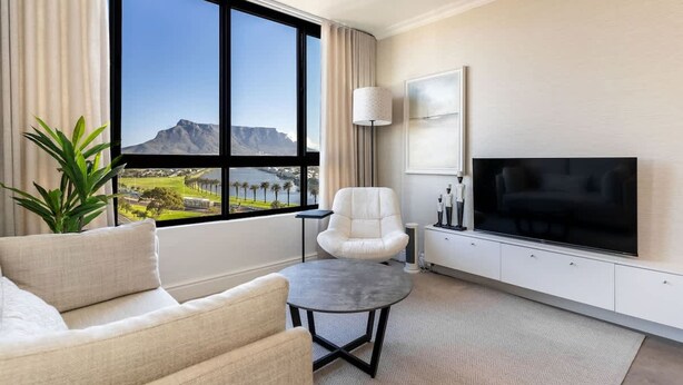 Habitación de hotel moderna con un amplio ventanal que ofrece vistas a la montaña, un televisor de pantalla plana, un sofá, una silla y una pequeña mesa.