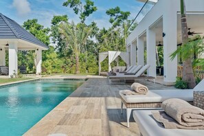 A modern outdoor pool area with lounge chairs and a covered seating area, surrounded by lush greenery. - Contemporary 5 BR Luxury Golf Villa w Large Pool (Punta Cana)