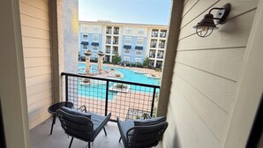 Un balcon agrémenté de deux fauteuils en osier et d’une petite table, d’où l’on peut admirer une piscine et un immeuble doté de nombreux balcons.