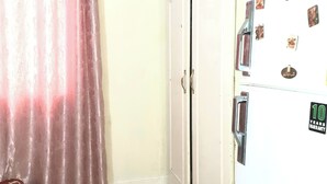 Une pièce avec une armoire blanche, une porte dotée d’une poignée, un réfrigérateur blanc orné d’aimants et un rideau rose.