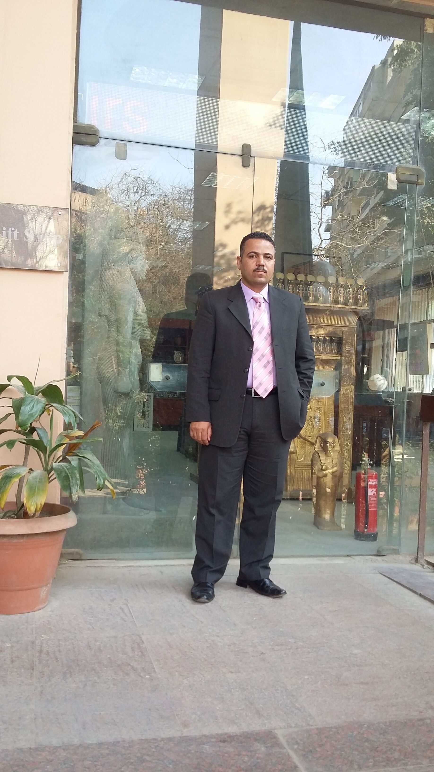A man standing in front of a building with a glass facade, a potted plant, and a fire extinguisher.