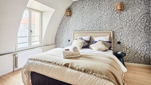 A bedroom with a sloped ceiling, a large bed with a wooden headboard, patterned wallpaper, and two wall-mounted lamps. - Flat Terrace Centre de Paris Bourse (Paris)