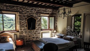 Ein rustikales Schlafzimmer mit Steinwänden, Holzbalkendecke, einem großen Bett mit vielen Kissen, einem kleinen Tisch mit Lampe und einem Sessel.