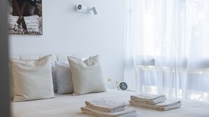 Una cama bien hecha con almohadas y toallas dobladas, un reloj en la pared y cortinas traslúcidas que dejan entrar luz natural.