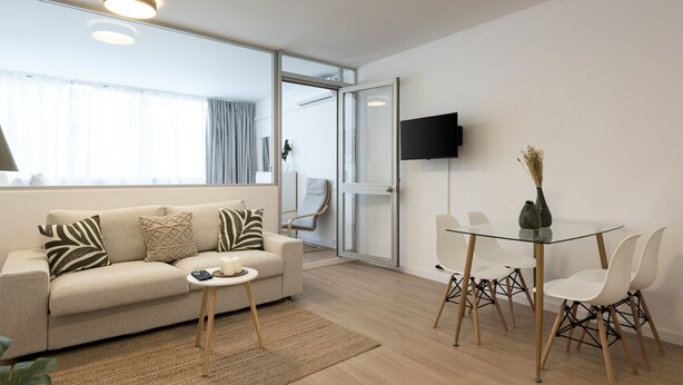 Una sala moderna con sofá, mesa de centro y mesa de comedor de vidrio con sillas. Un televisor está montado en la pared, y hay una puerta que da a otra habitación.