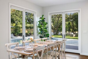 Uma sala de jantar com uma mesa posta para seis pessoas, janelas amplas e um vaso de planta.