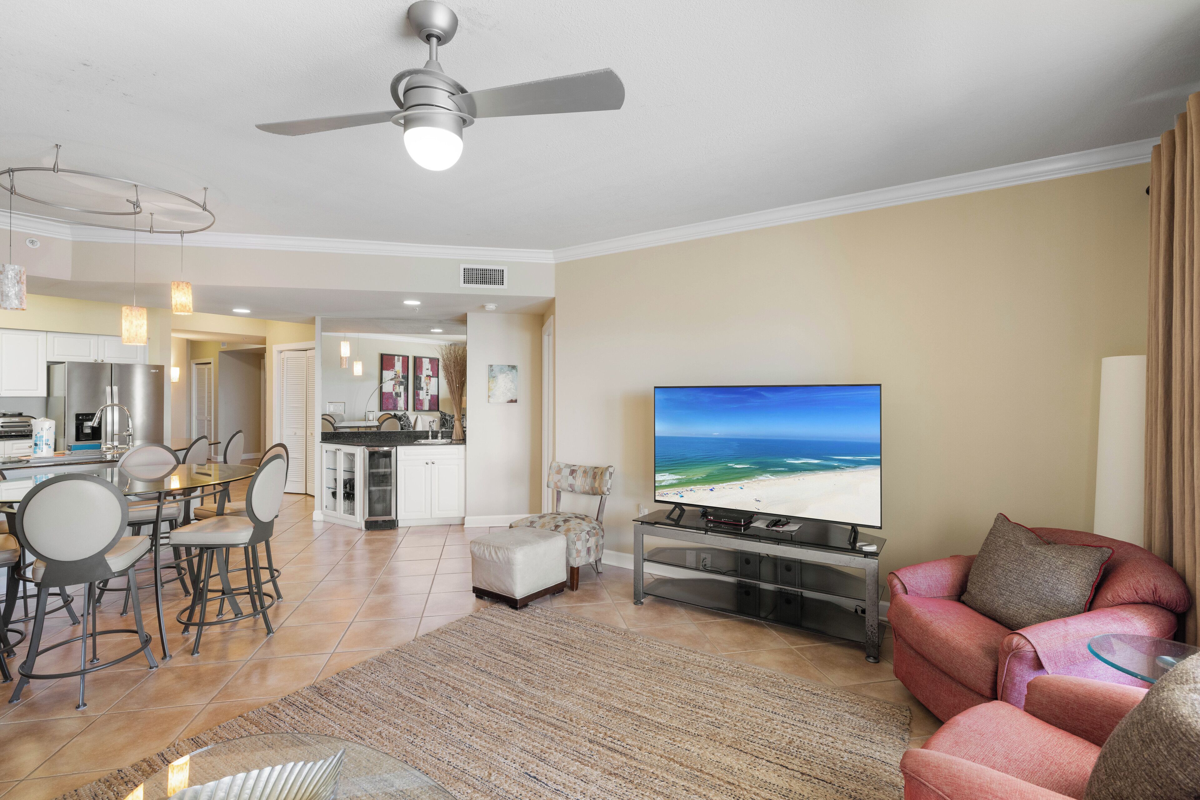 A spacious living room with a large flat-screen TV, a comfortable sofa, a dining area with chairs, and a kitchen in the background.