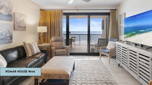 A spacious living room with a black leather sofa, a beige armchair, a small table with a lamp, and a large flat-screen TV on a wooden cabinet. A ceiling fan is visible, and the room has a large sliding glass door leading to a balcony with a view of the ocean.