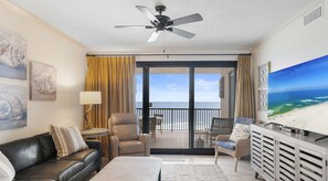 A spacious living room with a black leather sofa, a beige armchair, a small table with a lamp, and a large flat-screen TV on a wooden cabinet. A ceiling fan is visible, and the room has a large sliding glass door leading to a balcony with a view of the ocean.