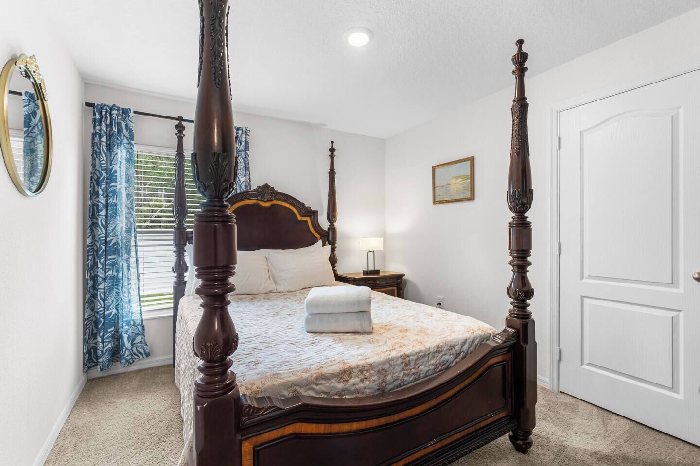 A four-poster bed with a floral bedspread, a bedside table with a lamp, and a large mirror on the wall.