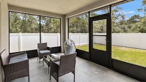 A covered patio area with wicker furniture, a grill, and a view of a white fence and trees.