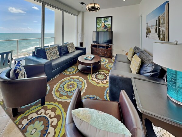 A modern living room with a large window overlooking the ocean, a flat-screen TV, and a colorful patterned rug. - Tranquility Beach | Oceanfront Condo (Galveston)