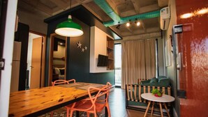 Un coin repas avec une table en bois et des chaises orange, un luminaire suspendu vert et une petite table ronde agrémentée d’une plante.