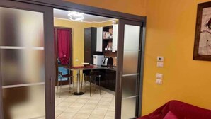 Ein Wohnzimmer mit einem roten Sofa, einer Glastür, die in einen Küchenbereich mit Esstisch und Stühlen führt, und einem roten Vorhang.