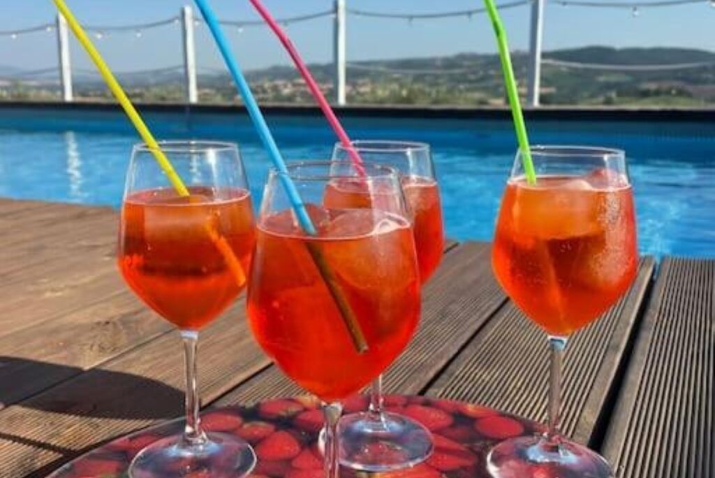 Three glasses of red beverage with colorful straws on a wooden table by a pool with a bridge in the background.