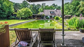 Una terraza de madera con dos sillones de descanso orientados hacia un estanque y una casa al fondo.