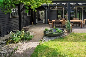 A covered patio with a brick pathway, wooden furniture, and a garden bed with plants. - Attractive villa in the middle of the woods, pet-friendly with a swimming pond (Ede)