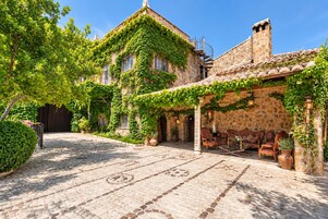 A courtyard with stone flooring, a covered seating area with chairs, and a building with stone walls and a tiled roof.