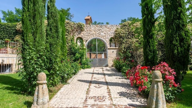 A stone pathway leading to a rustic building with a metal gate, surrounded by lush greenery and flowering plants. - PALACIO ARUDECAS (Cañada de Calatrava)
