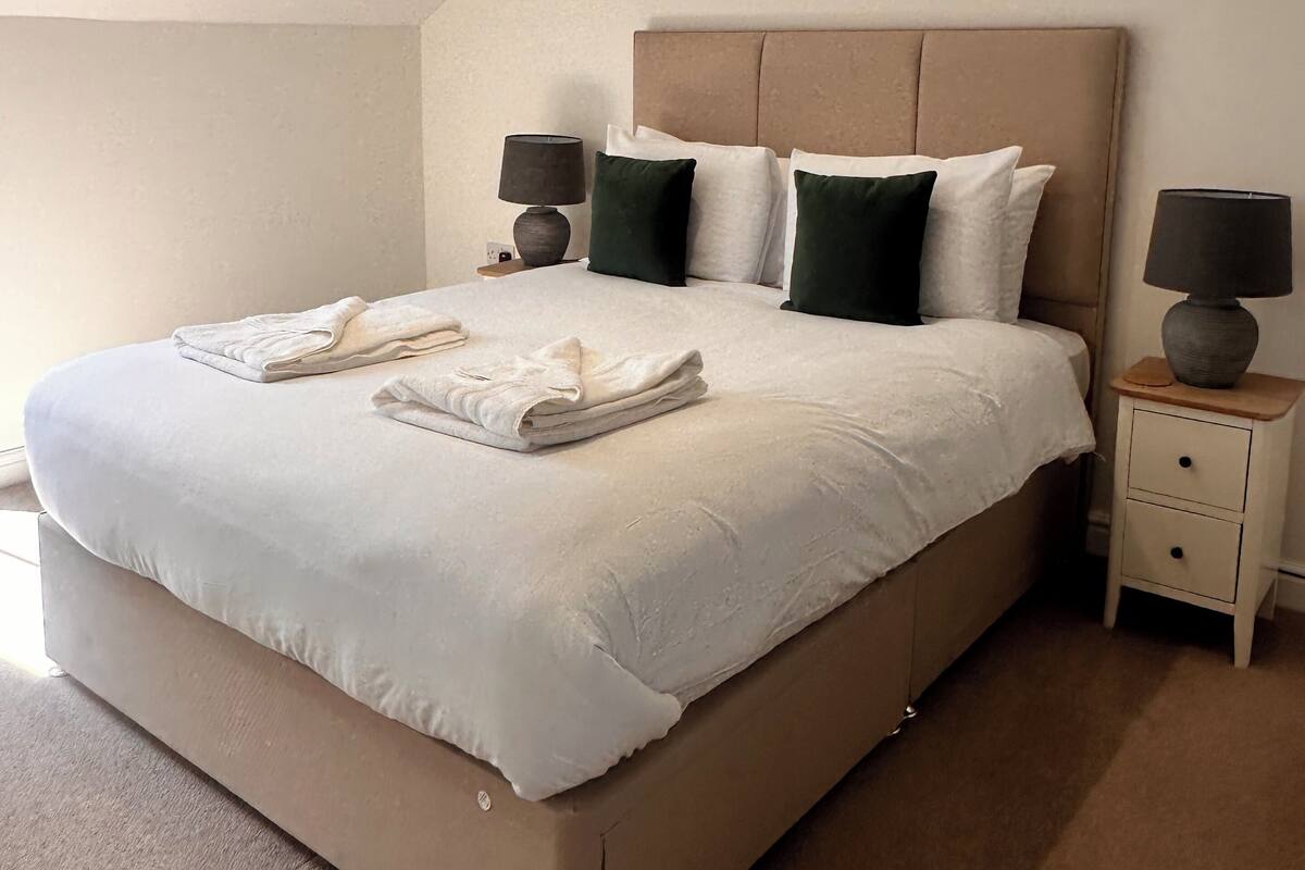 A neatly made bed with white linens and two towels, a headboard, two bedside tables with lamps, and a sloped ceiling.