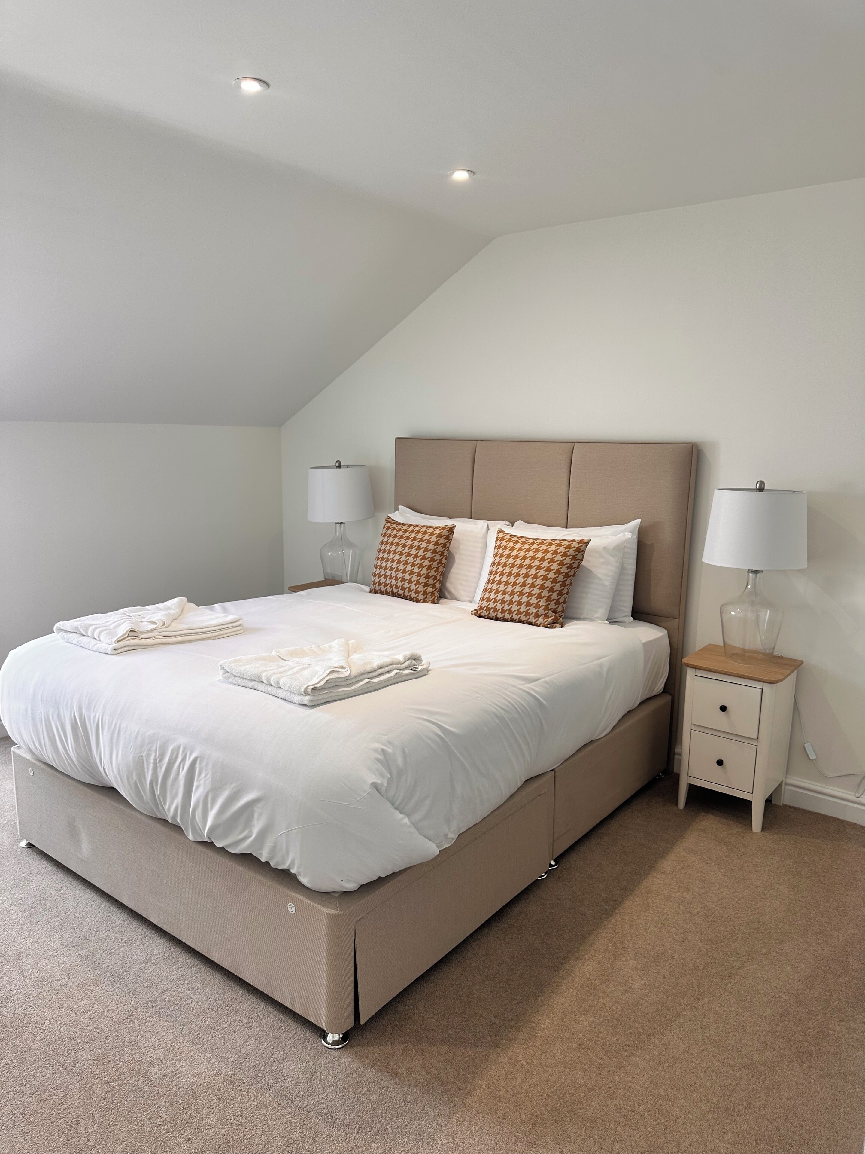 A neatly made bed with white linens and patterned pillows, a bedside table with a lamp, and a carpeted floor.
